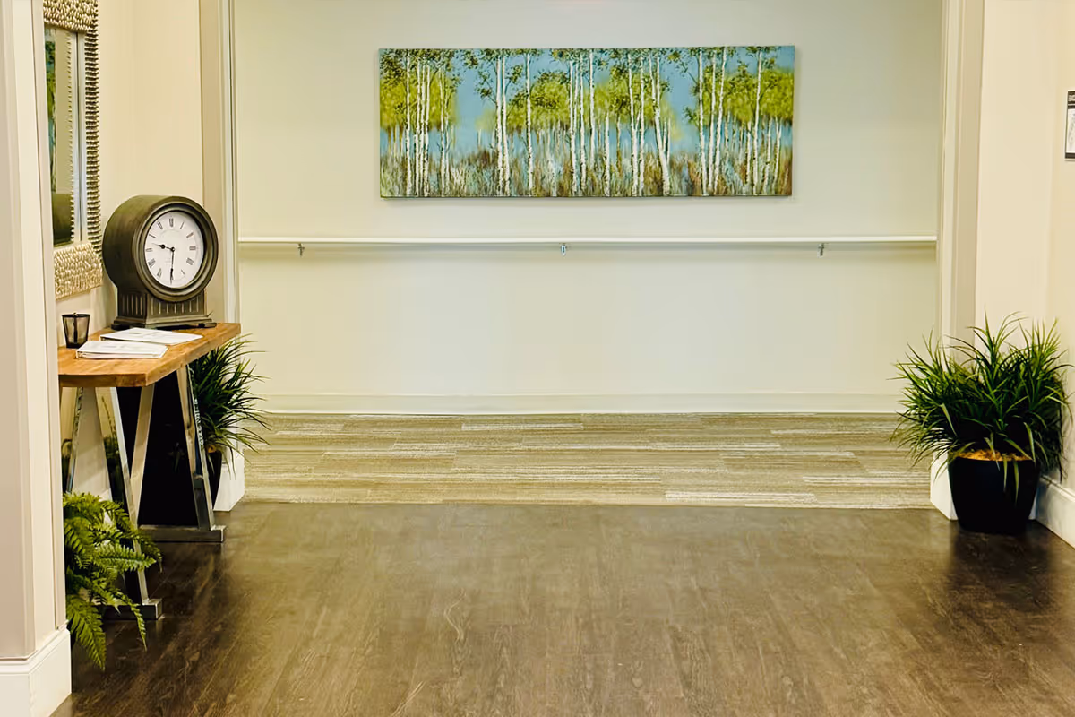 A hallway with wooden flooring and light-colored walls featuring a large painting of trees. There is a wooden console table on the left with a round clock, a candle, and some papers. Green potted plants are placed on both sides of the hallway.