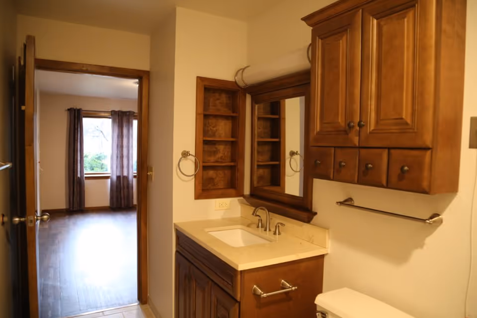 Interior view of a bathroom with wooden cabinets, a sink with a countertop, a mirror, and a towel rack. The bathroom door is open, showing a room with wooden flooring and a window with curtains.