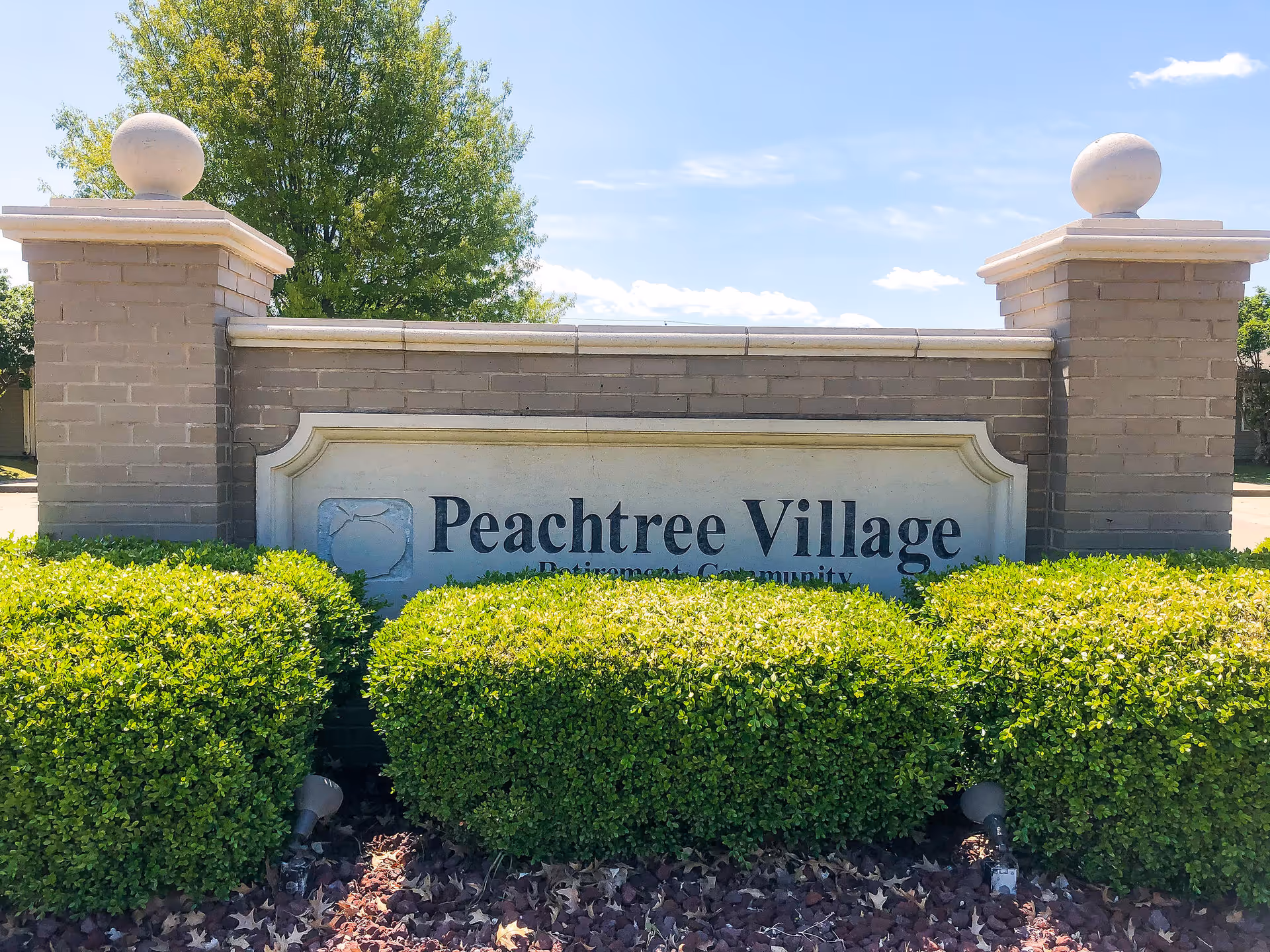 A brick and stone sign for Peachtree Village Retirement Community surrounded by green bushes with a tree and blue sky in the background.