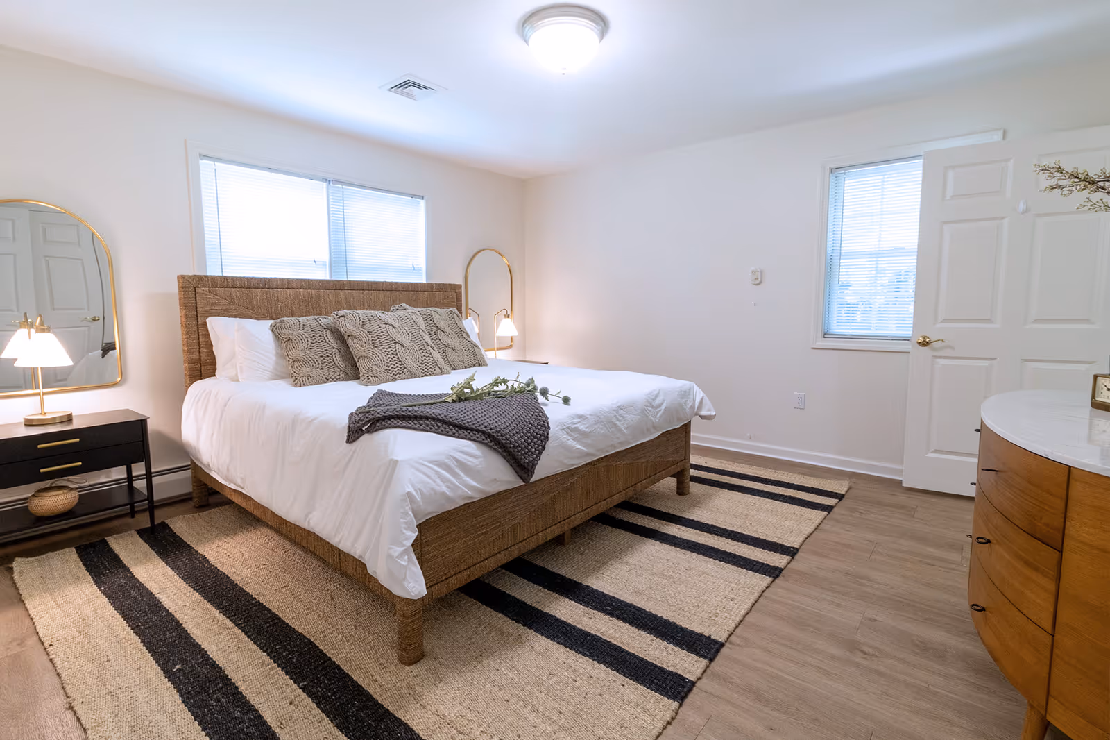 Sunlit bedroom with a woven bed frame, white bedding and pillows, two bedside lamps, a striped rug, and a wooden dresser.