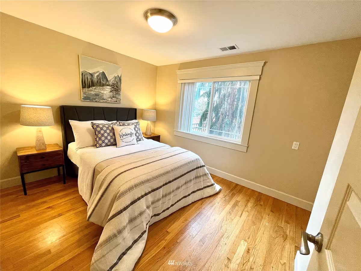 A cozy bedroom with a neatly made bed featuring white and patterned pillows, a striped blanket, two wooden nightstands with lamps, a window with a view of trees, and a painting of mountains on the wall above the bed.