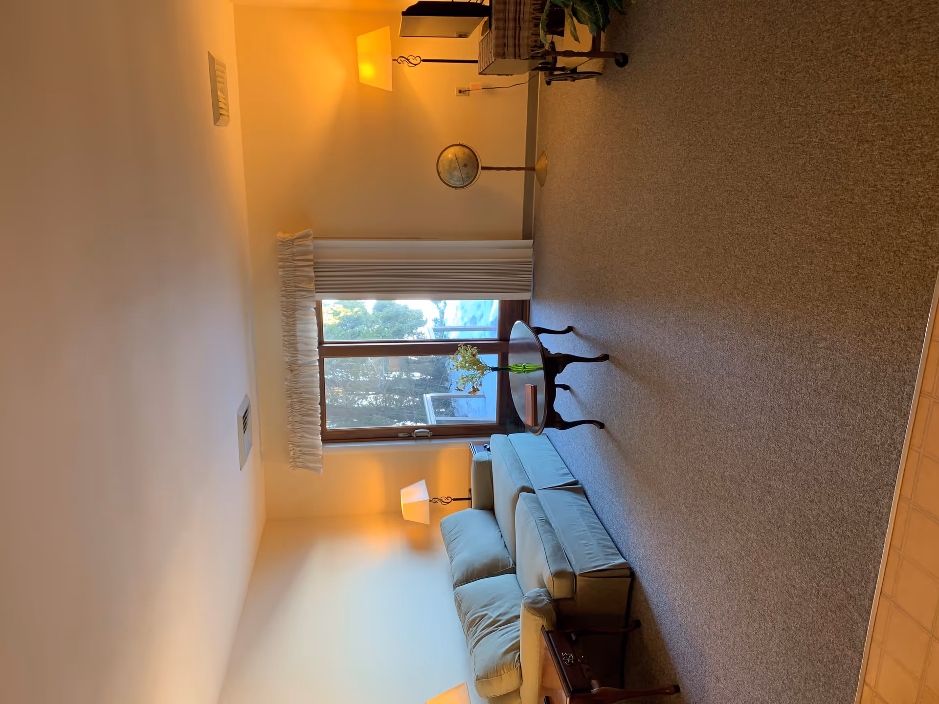A cozy living room with a light blue sofa, a wooden coffee table with a glass vase and flowers, a floor lamp, a globe on a stand, and a large window with white curtains letting in natural light.