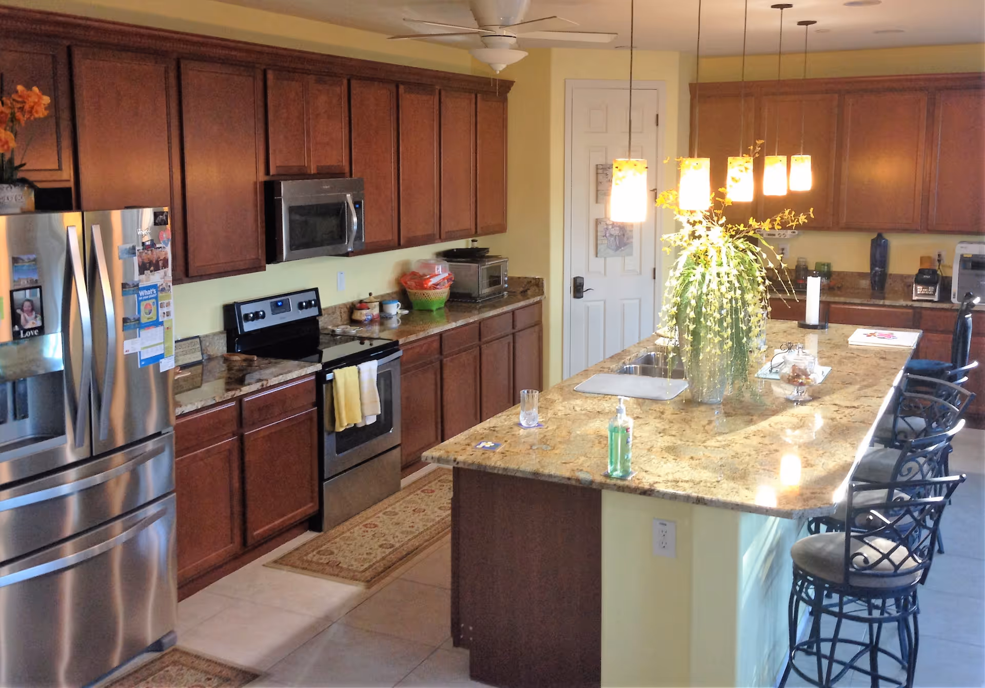 A spacious kitchen with wooden cabinets, a stainless steel refrigerator, stove, and microwave. There is a large granite island in the center with a sink, a vase with green plants, and several hanging pendant lights above. Three bar stools are placed along one side of the island.