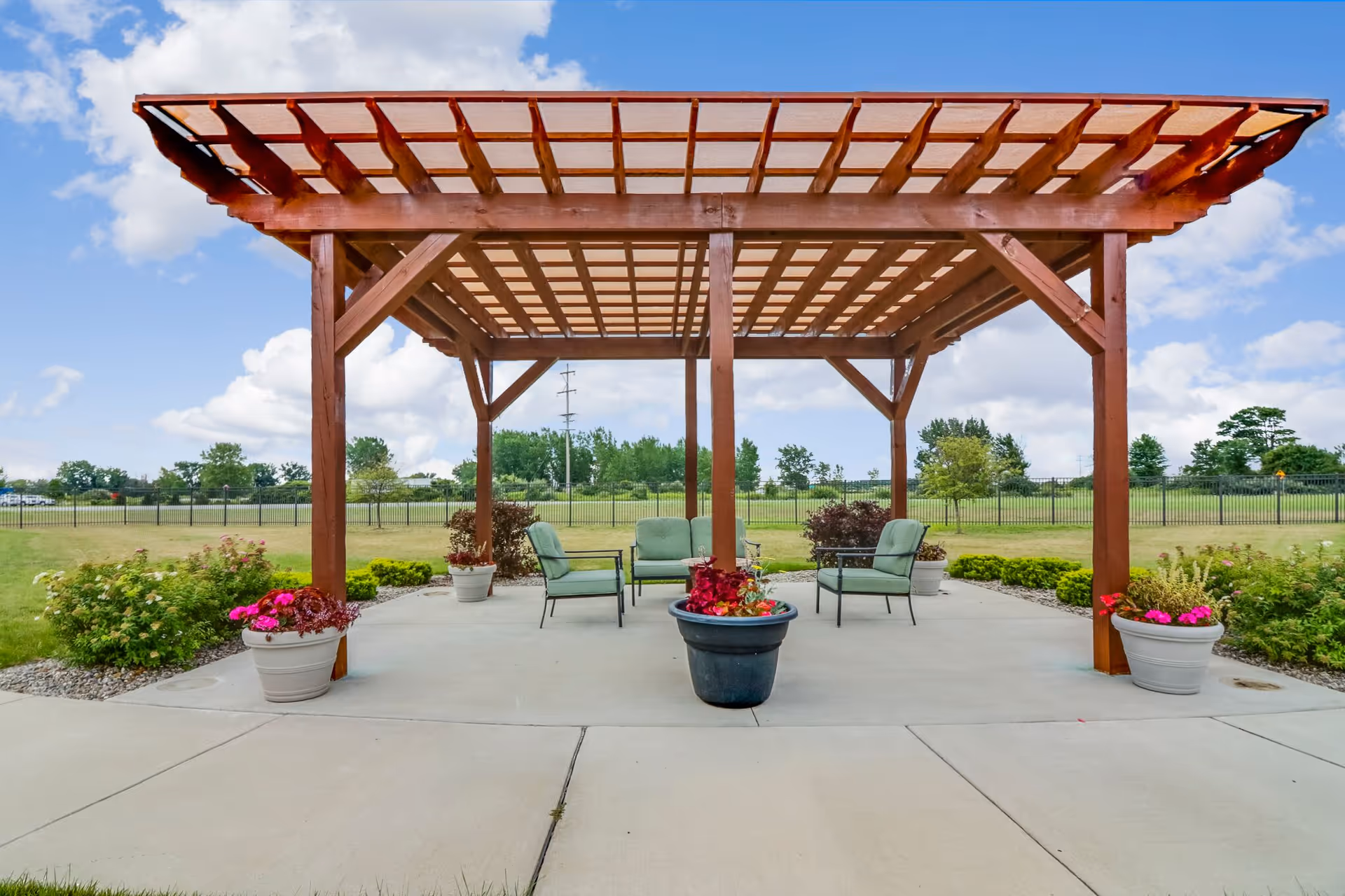 Outdoor seating area with a wooden pergola providing partial shade over a concrete patio. The area includes cushioned chairs and a loveseat arranged around a central planter with flowers. Additional flower pots and landscaped greenery surround the patio, with a grassy field and trees in the background under a partly cloudy sky.