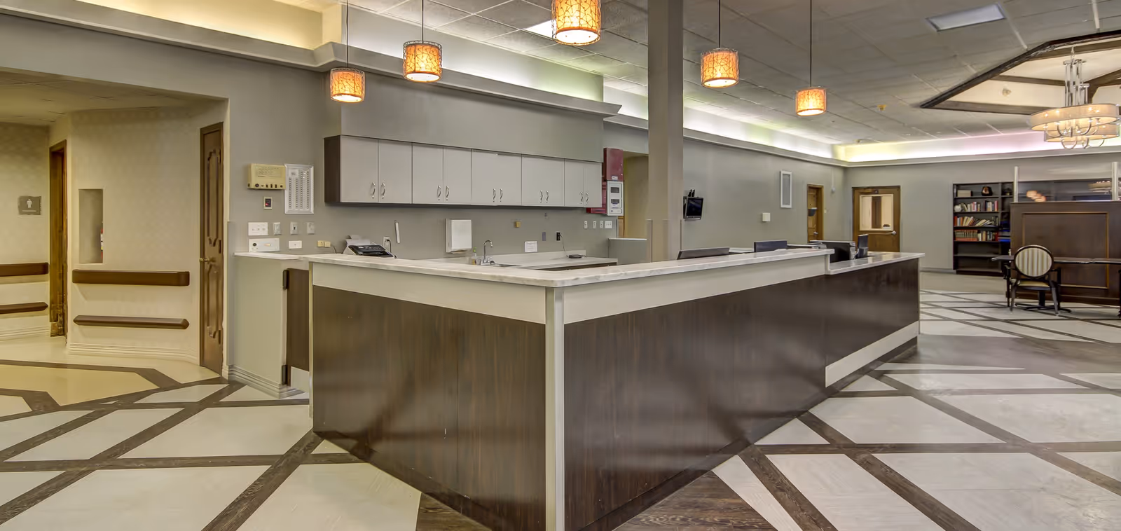 Interior view of a senior living facility reception area with a large dark wood reception desk, overhead pendant lights, and a patterned floor. The background shows a seating area with chairs and bookshelves.