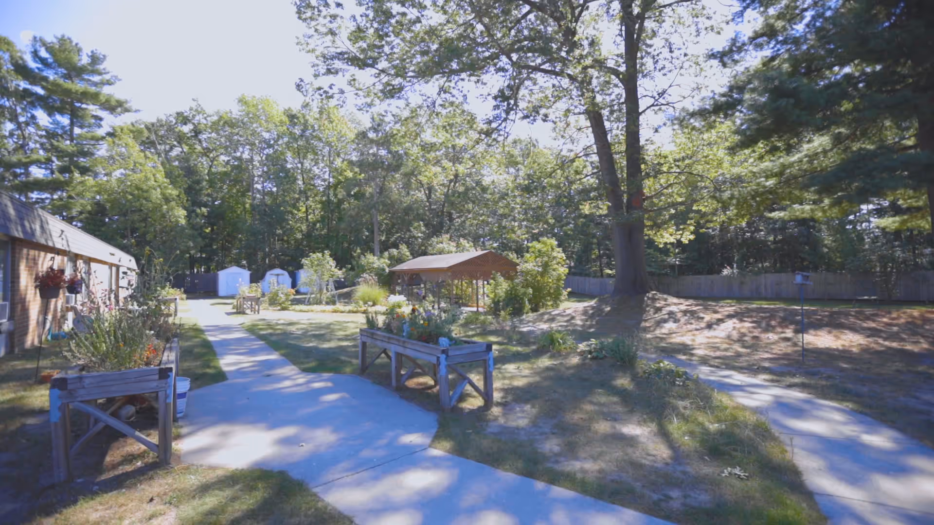 Outdoor garden area at Medilodge of GTC with paved walkways, raised garden beds, a wooden gazebo, and surrounded by trees and greenery under a clear sky.