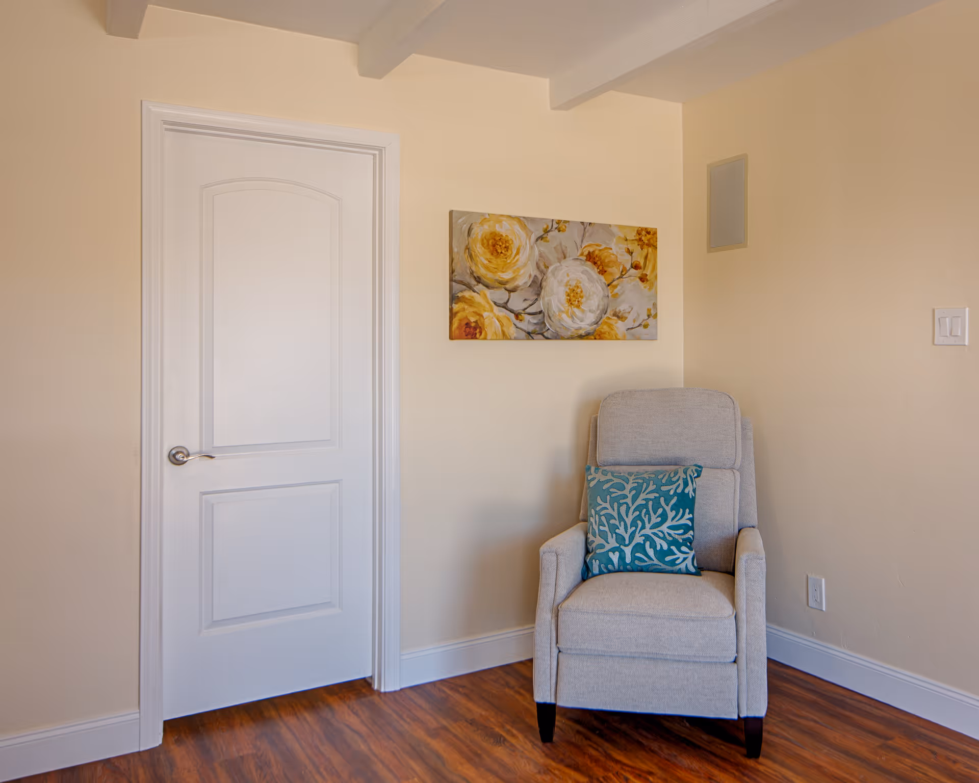 A beige upholstered armchair with a teal patterned pillow sits beside a closed white door beneath a floral painting in a softly lit room.