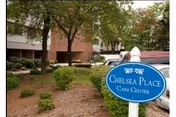 Outdoor view of Chelsea Place Care Center showing a landscaped area with bushes and trees in front of a multi-story building. A blue sign with white text reads 'Chelsea Place Care Center.'