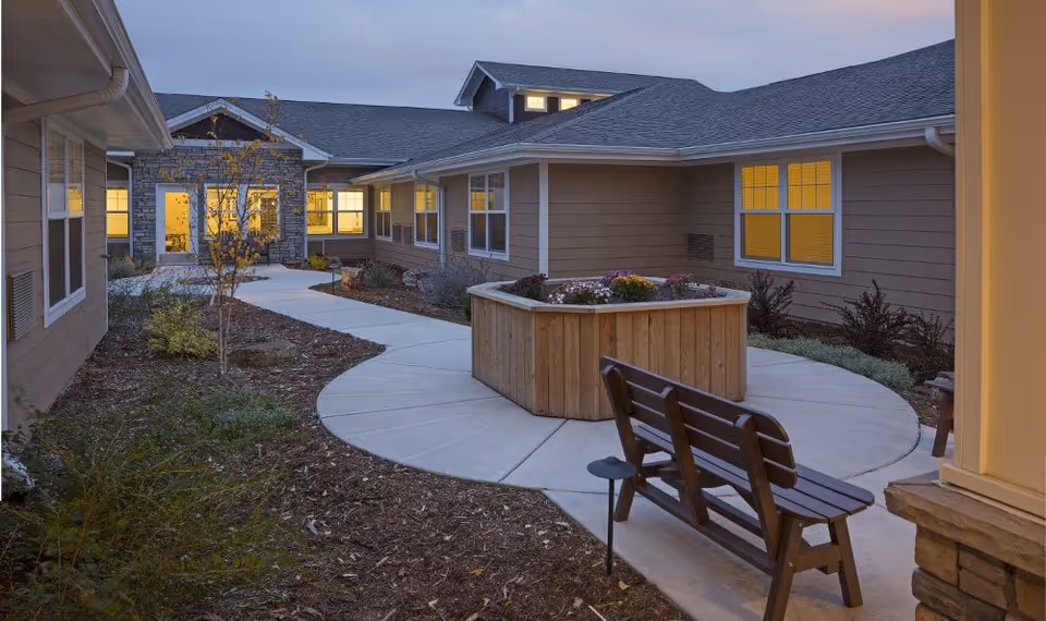Outdoor courtyard area at dusk with a curved concrete walkway, a wooden bench, and a raised wooden planter filled with flowers. The surrounding buildings have beige siding and multiple windows with warm interior lighting.