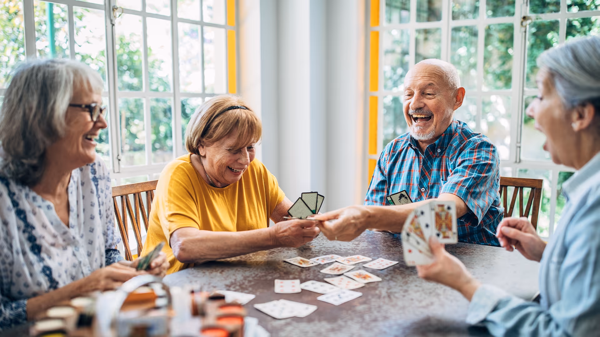 Four elderly people sitting around a table playing cards and laughing together in a bright room with large windows.
