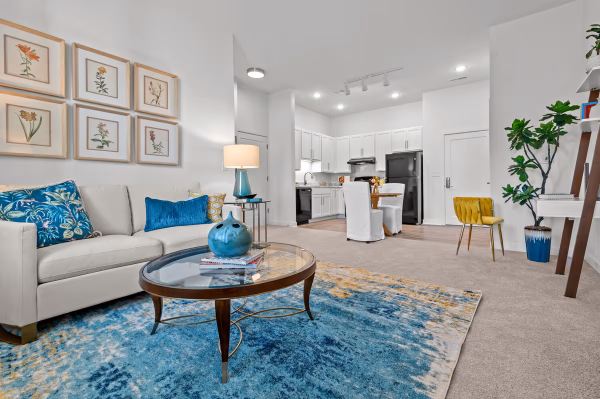 A bright and modern living area with a beige sofa adorned with blue and yellow pillows, a glass coffee table with a blue decorative vase, and a blue and yellow patterned rug. The background shows a white kitchen with black appliances, a small dining table with white slipcovered chairs, a yellow chair, and a tall green plant in a blue pot. The walls are white with framed botanical prints above the sofa.