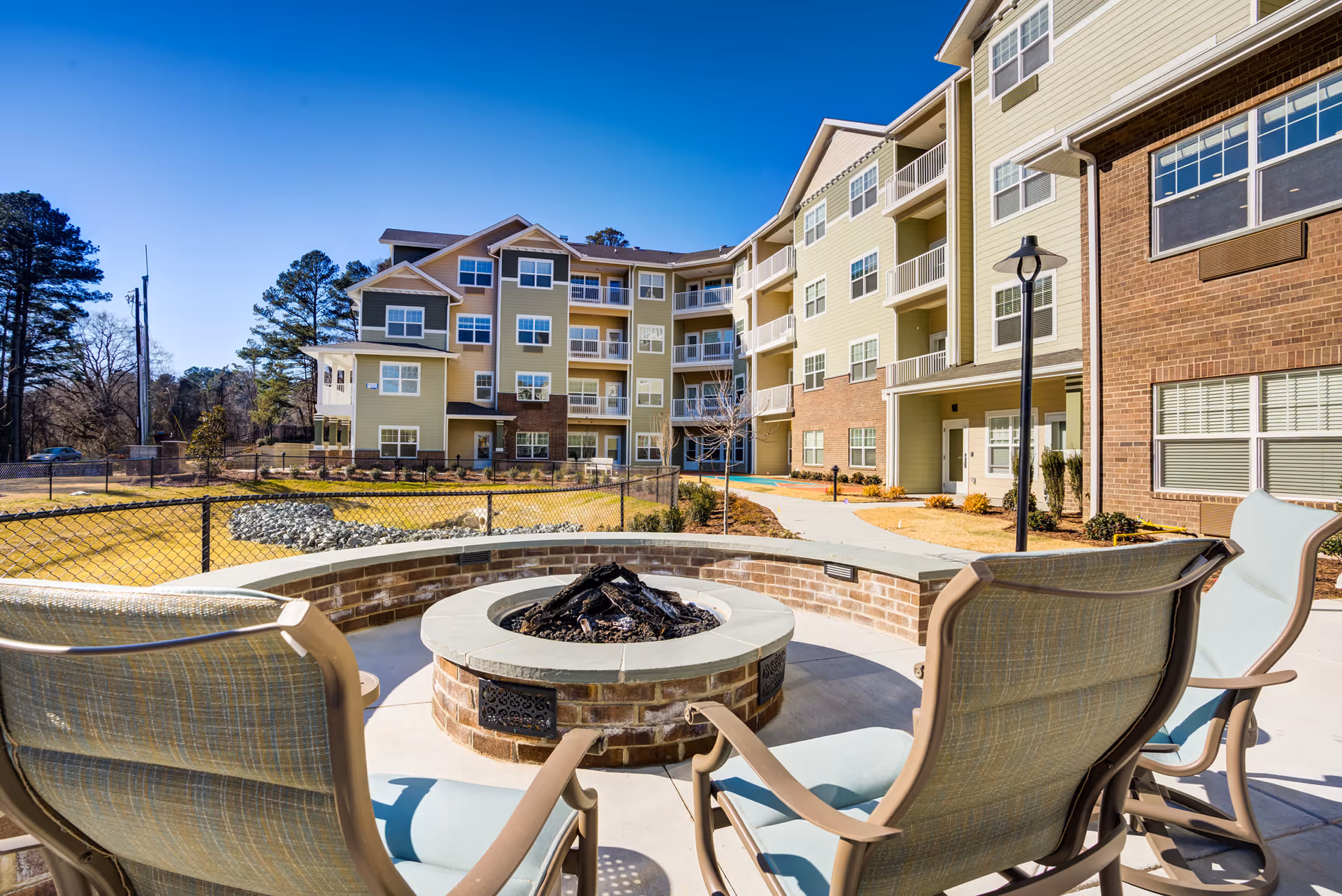 Outdoor seating area with cushioned chairs around a circular brick fire pit, set in front of a multi-story residential building with balconies and large windows under a clear blue sky.