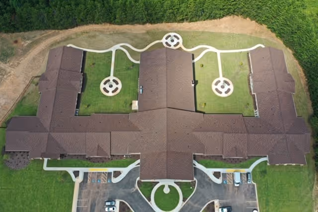 Aerial view of an assisted living building with a cross-shaped roof, landscaped lawns, circular walkways, and a parking lot.