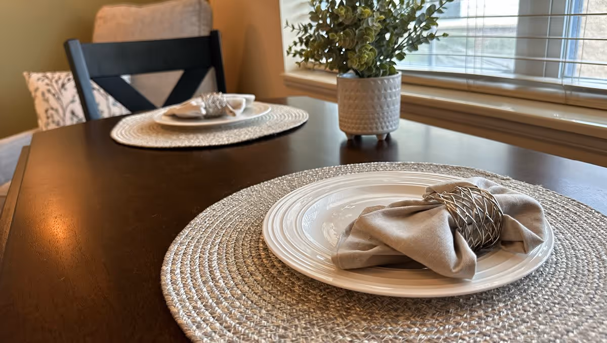Close-up view of a dark wooden dining table set with woven placemats, white plates, and beige cloth napkins held by silver napkin rings. A potted plant sits near a window with blinds in the background.