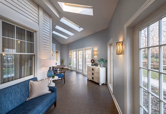 A bright and airy interior hallway with skylights and large windows on one side, featuring a blue sofa with a beige pillow, a small round table with a lamp, a wooden table with chairs, and a white chest of drawers decorated with plants and ornaments.