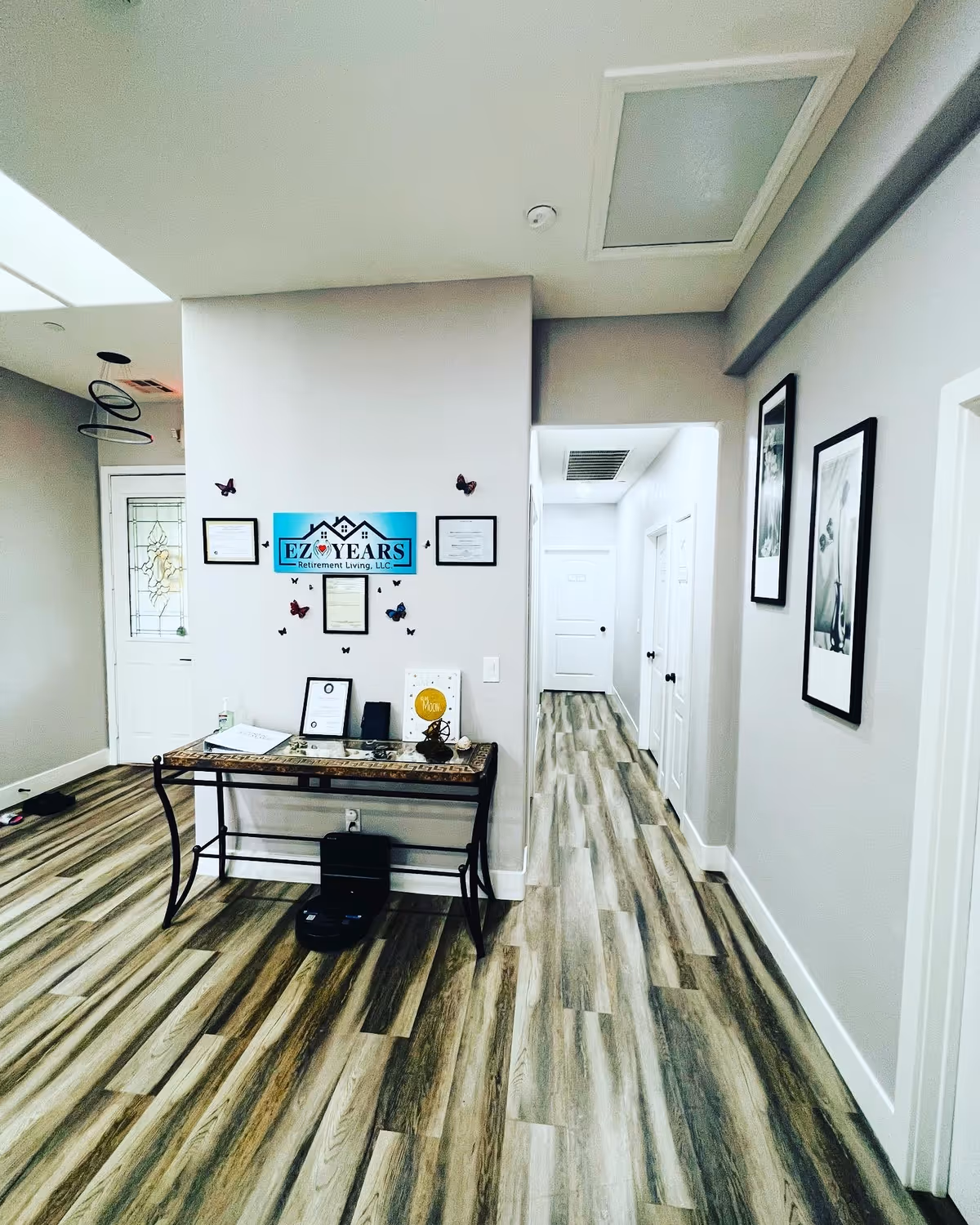 Interior view of a hallway in EZ Years Retirement Living facility with wood-patterned flooring, light gray walls, a decorative table with framed certificates and a sign on the wall, butterfly wall decorations, and framed pictures on the right wall.
