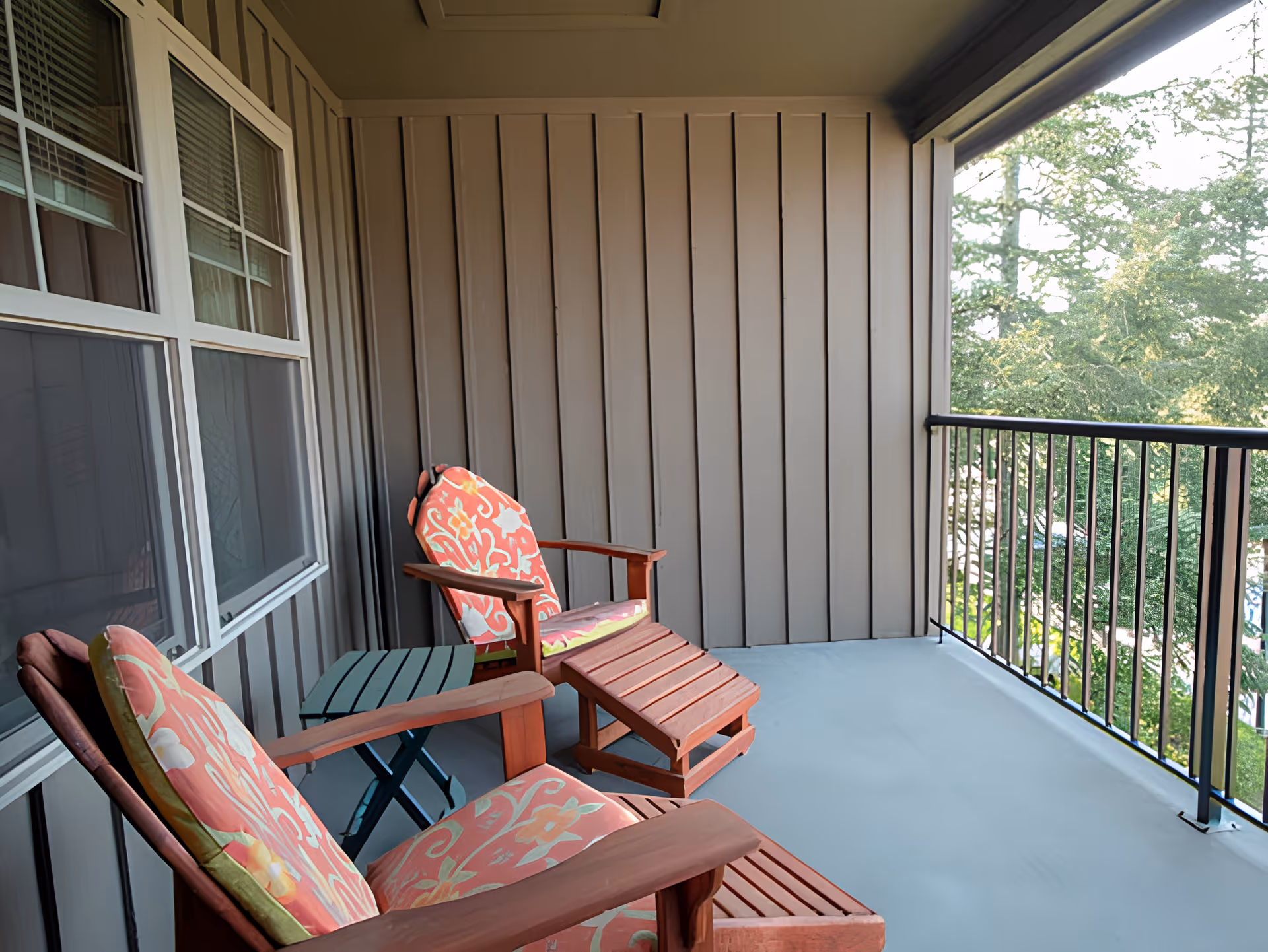 A covered balcony with two wooden lounge chairs featuring floral cushions and a small green side table between them. The balcony has a metal railing and overlooks trees and a parking area.