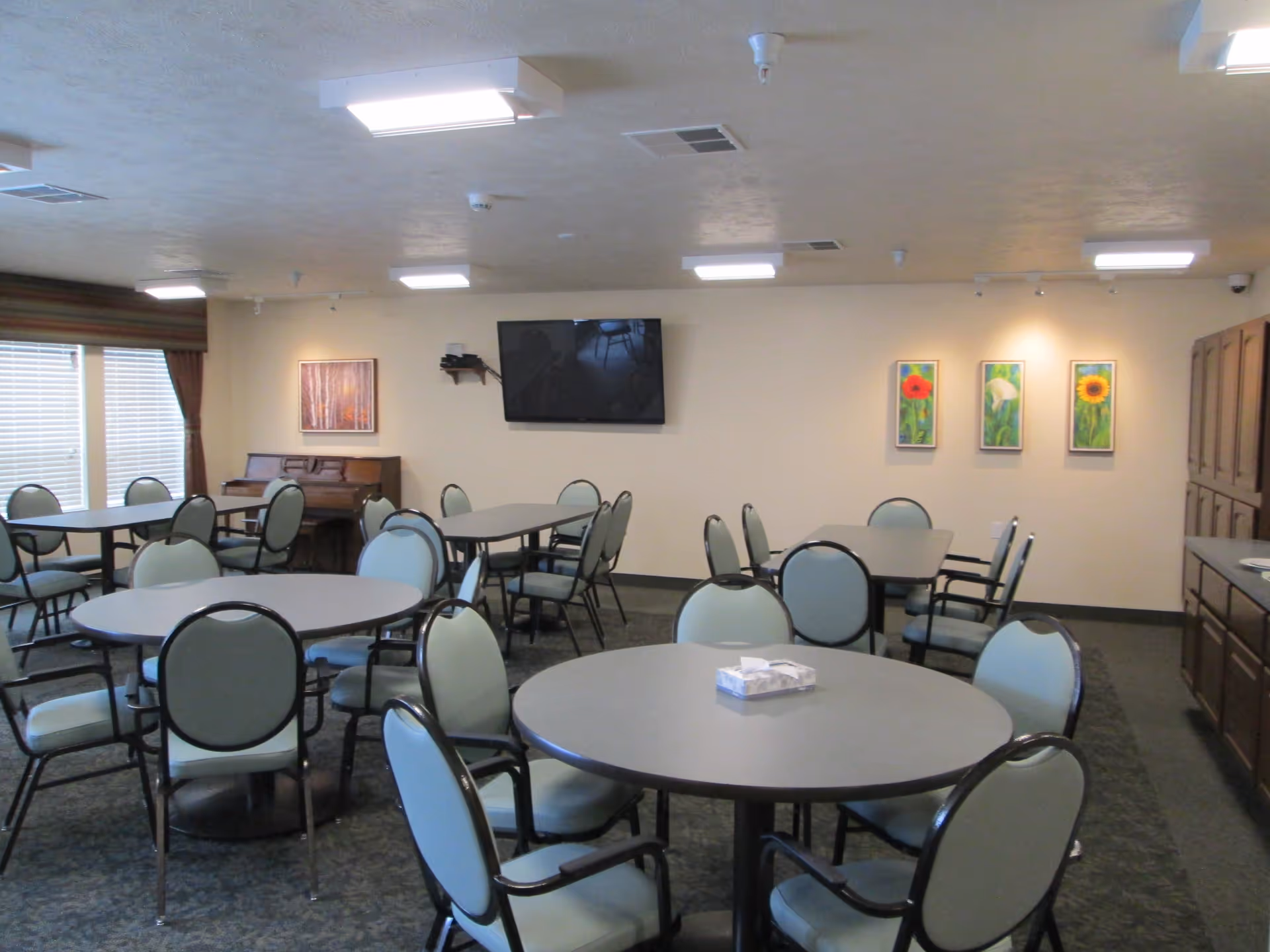 Communal dining room with round tables and green cushioned chairs, a wall-mounted TV, piano, and floral artwork on the wall.