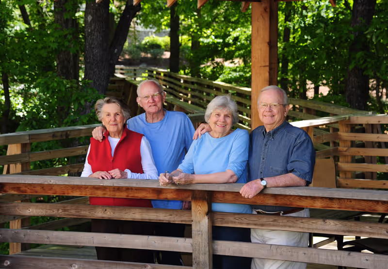 Four elderly people standing together on a wooden deck outdoors, surrounded by green trees. They are smiling and appear to be enjoying the natural setting.