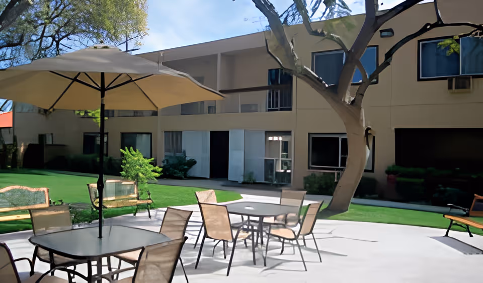 Outdoor courtyard with patio tables, chairs, umbrellas, benches, a large tree, and a two-story building in the background.
