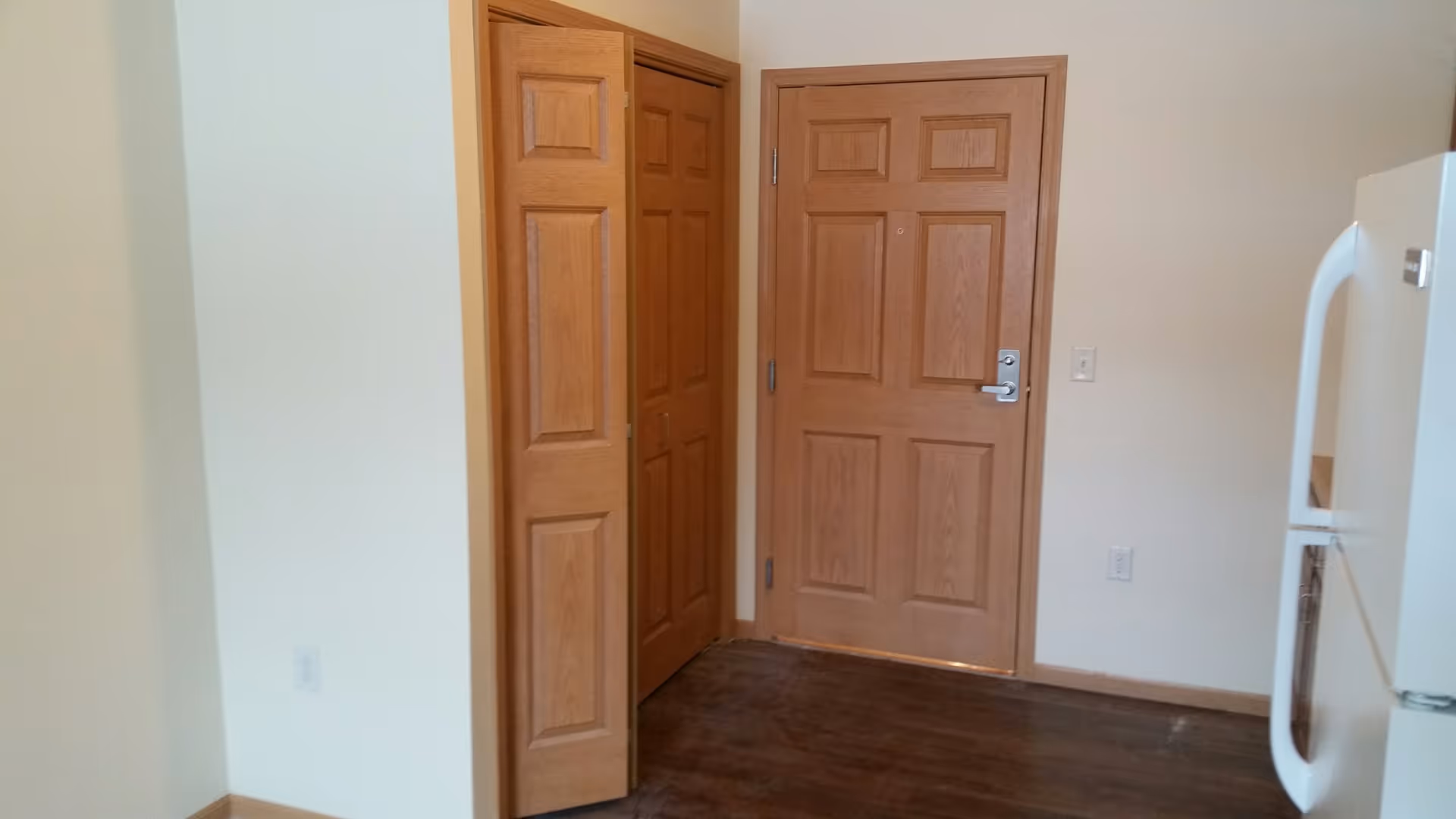 Interior view of a room corner showing a closed wooden entrance door, a partially open wooden closet door, light-colored walls, dark wood flooring, and a white refrigerator on the right side.