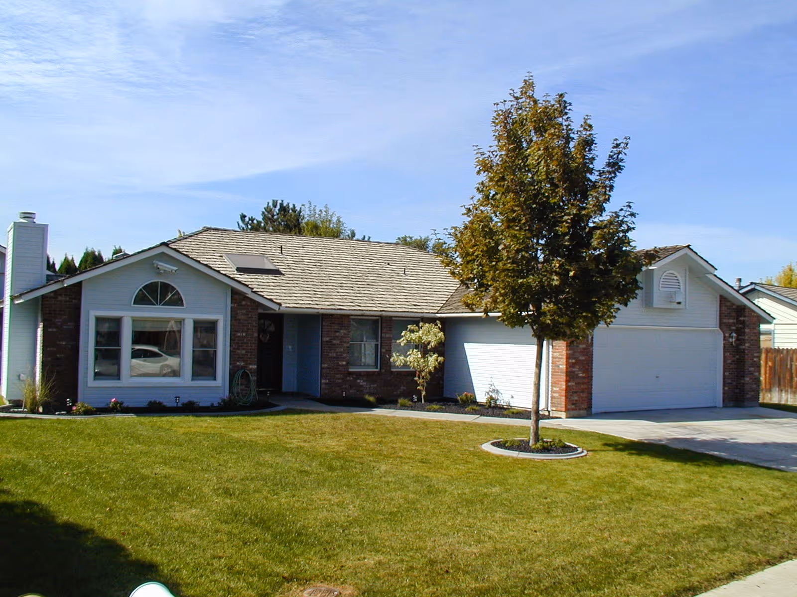 Single-story residential building with a brick and white exterior, a large front window, a two-car garage, a tree in the front yard, and a well-maintained green lawn under a clear blue sky.