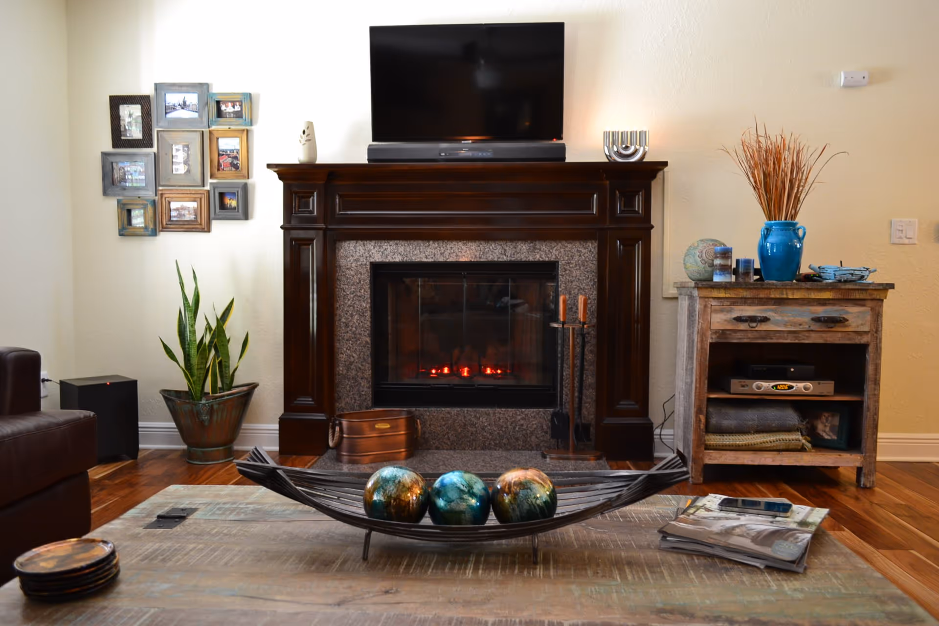 A cozy living room with a dark wooden fireplace mantel, a flat-screen TV mounted above it, and a soundbar below the TV. On the mantel are decorative items including a white vase and a silver candle holder. To the right of the fireplace is a rustic wooden side table with a blue vase holding dried branches, a globe, candles, and other small items. To the left is a green potted plant and a collage of framed photos on the wall. In front of the fireplace is a wooden coffee table with a decorative metal bowl holding three colorful spheres, a stack of magazines, and a smartphone.
