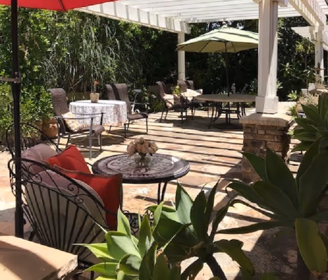Outdoor patio with tables, chairs, umbrellas, and potted plants under a pergola.