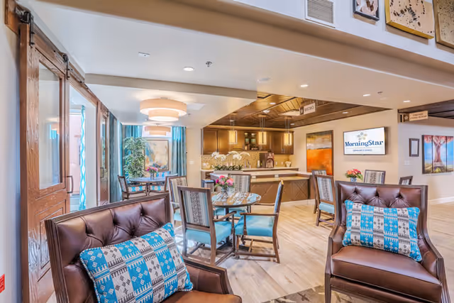 A bright and welcoming interior common area in MorningStar Assisted Living & Memory Care of Santa Fe featuring comfortable brown leather chairs with patterned blue and white pillows, round tables with chairs, a kitchen area with dark wood cabinets, and decorative artwork on the walls. The space is well-lit with ceiling lights and natural light from large windows with blue curtains.