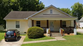 Single-story beige house with a front porch, steps leading up to the porch, a bush in front, a black car parked in the driveway, and green grass lawn under a clear blue sky.