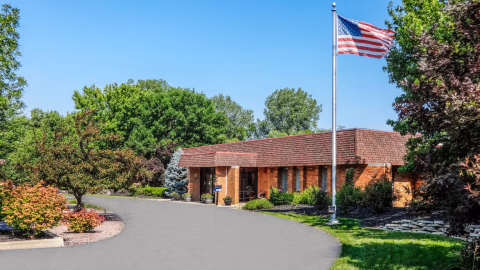 Single-story brick facility entrance with an American flag on a flagpole, circular driveway, and landscaped trees and shrubs.