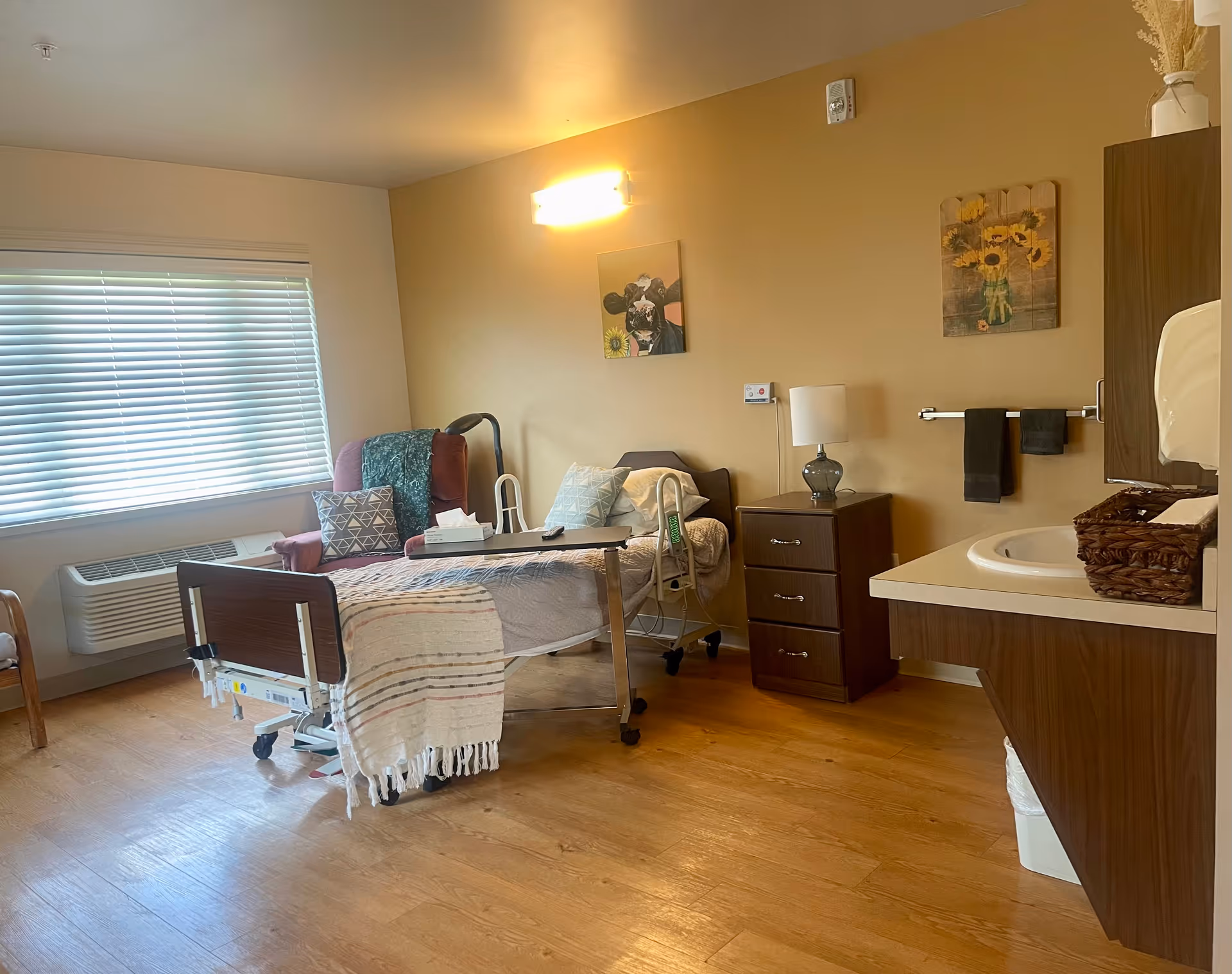 A well-lit senior care facility room with a hospital bed covered with a blanket and pillows, a maroon recliner chair with a pillow and blanket, a wooden nightstand with a lamp, a wall-mounted light, two paintings of sunflowers and a dog, a sink with a countertop and baskets, and a window with blinds.