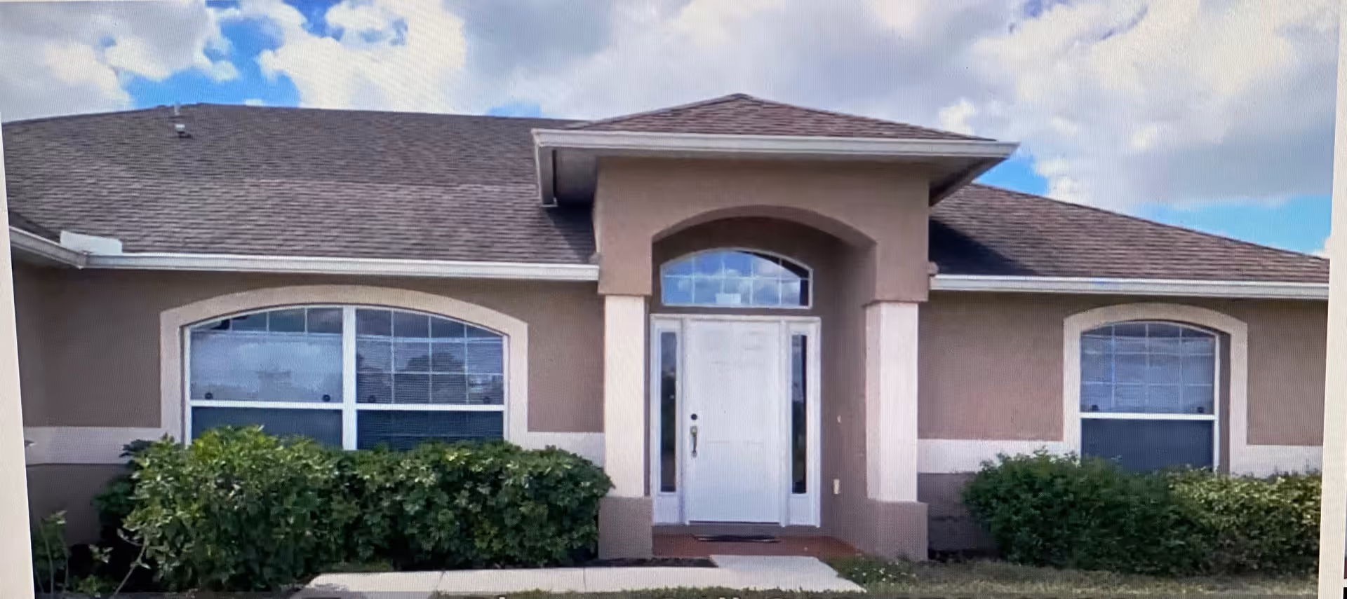 Front exterior view of a single-story building with a central white door flanked by two large windows. The building has a beige facade with a brown roof and is surrounded by green bushes and a small lawn under a partly cloudy sky.
