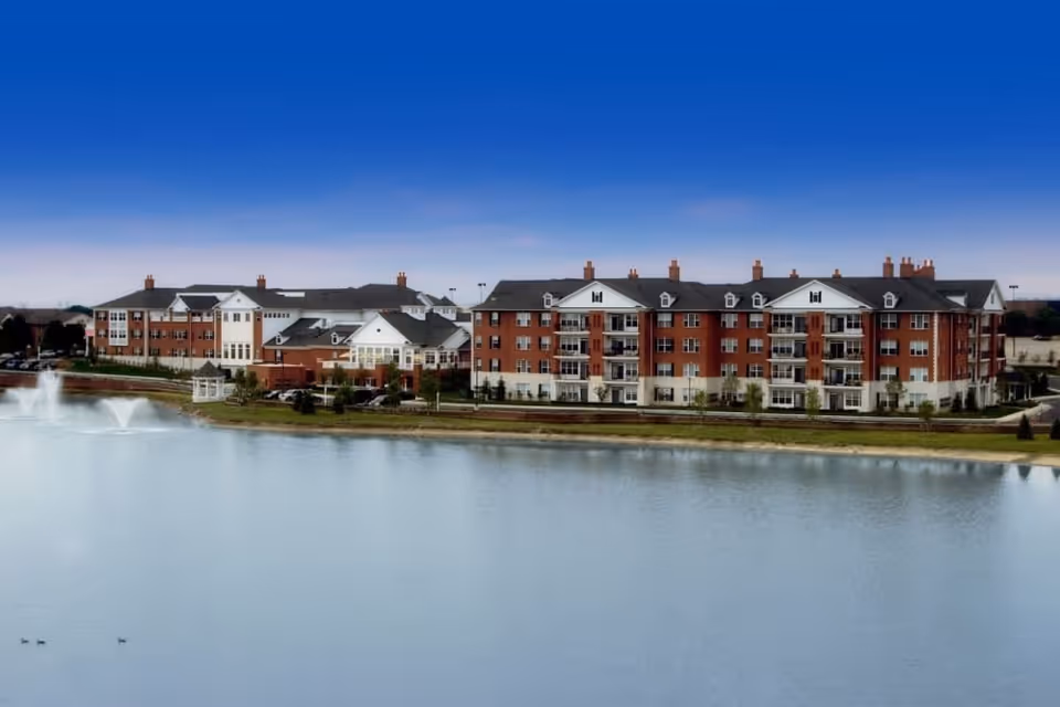 A large senior living facility named Waltonwood Lakeside situated beside a calm lake with two water fountains. The building is multi-story with red brick and white accents, featuring multiple windows and balconies. The sky above is clear and blue.