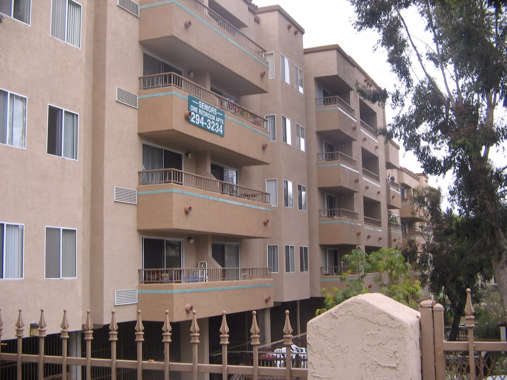 Exterior view of a multi-story senior living apartment building with balconies and a sign advertising one-bedroom apartments for seniors with a contact phone number. The building is beige with turquoise trim and is partially obscured by trees and a decorative metal fence in the foreground.