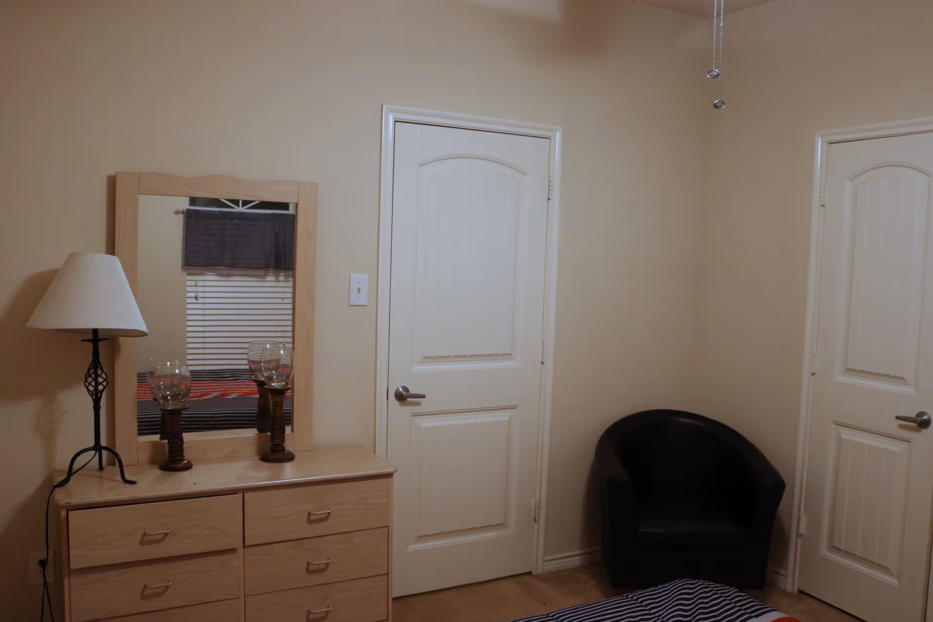 A bedroom corner with a light wooden dresser topped with a lamp and two decorative glass candle holders. Above the dresser is a mirror reflecting a window with closed blinds and a dark valance. To the right, there are two closed white doors and a black armchair in the corner. The room has beige walls and carpeted floor.