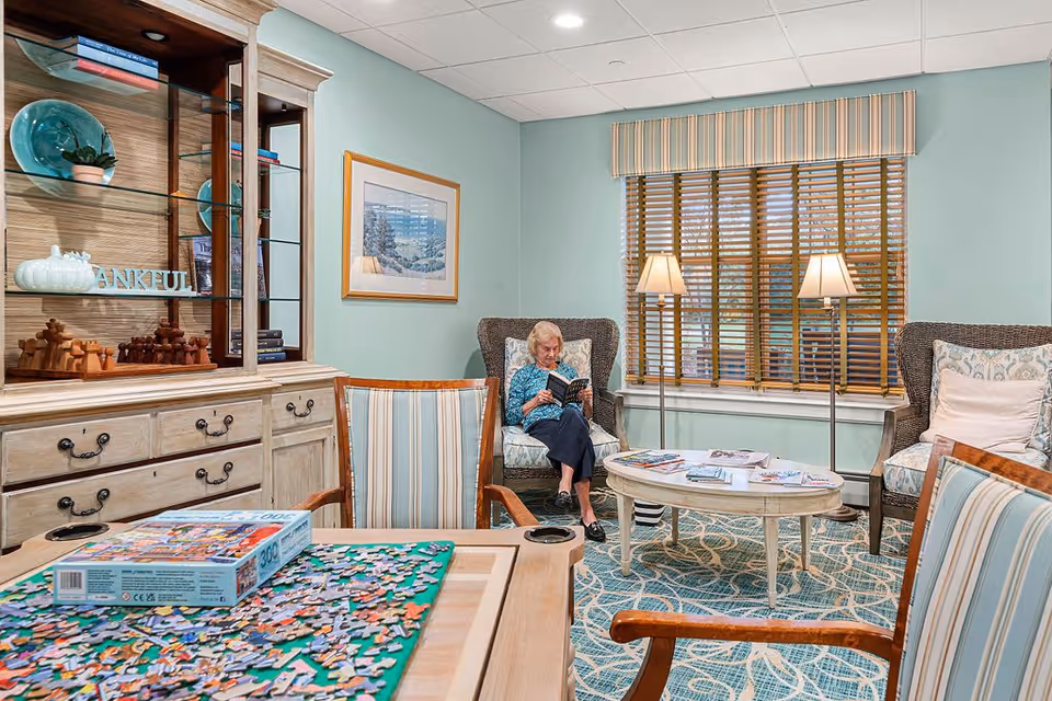 A cozy living room area with a woman sitting in a cushioned armchair reading a book. The room features a wooden cabinet with glass shelves displaying decorative items, a round coffee table with magazines, two floor lamps, a large window with wooden blinds and a striped valance, and a table in the foreground with a partially completed jigsaw puzzle and striped chairs.