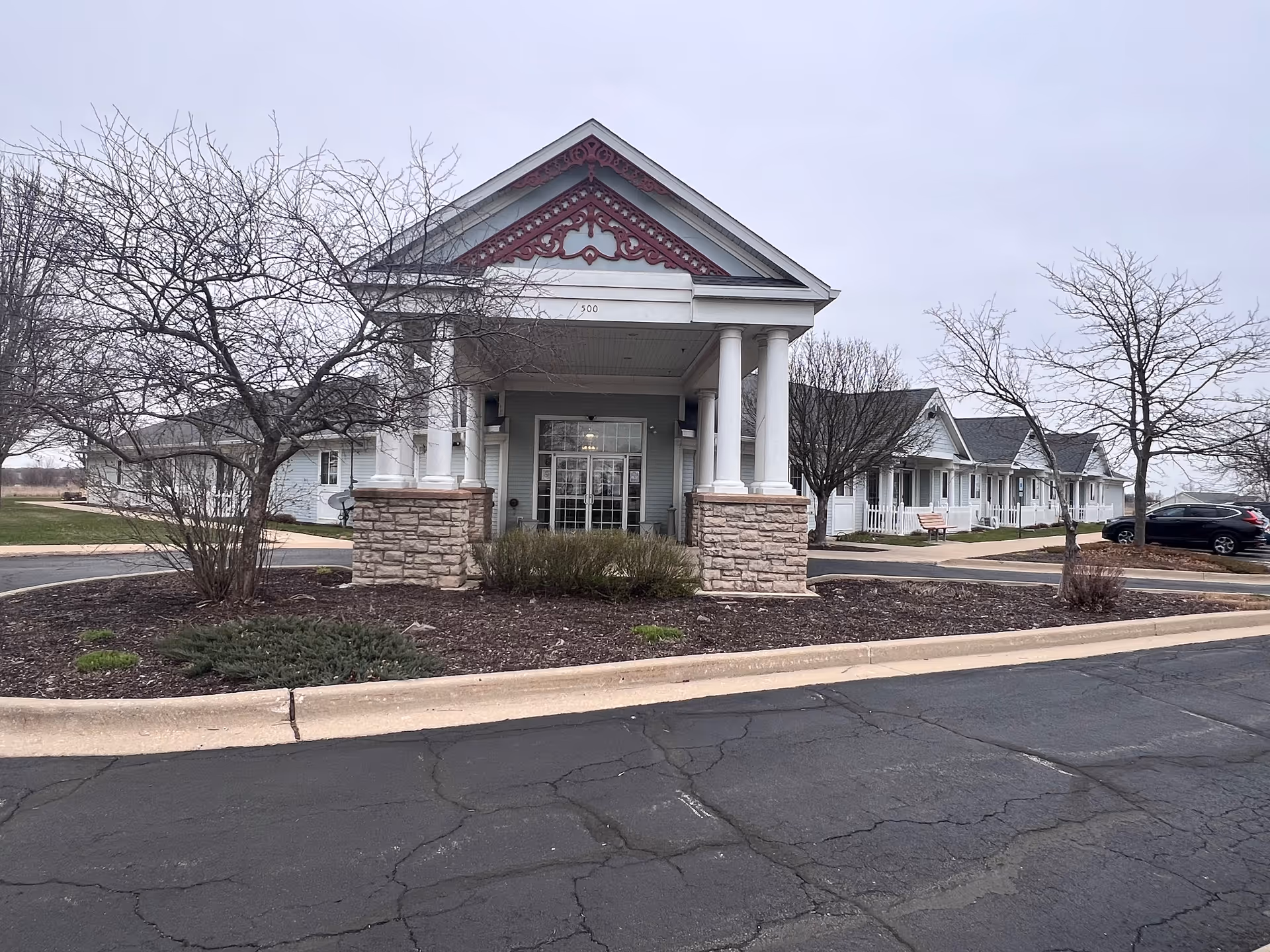Front exterior view of Dimensions Living Winnebago facility showing a covered entrance with white columns and stone bases, leafless trees, a parking area with cars, and a cloudy sky.