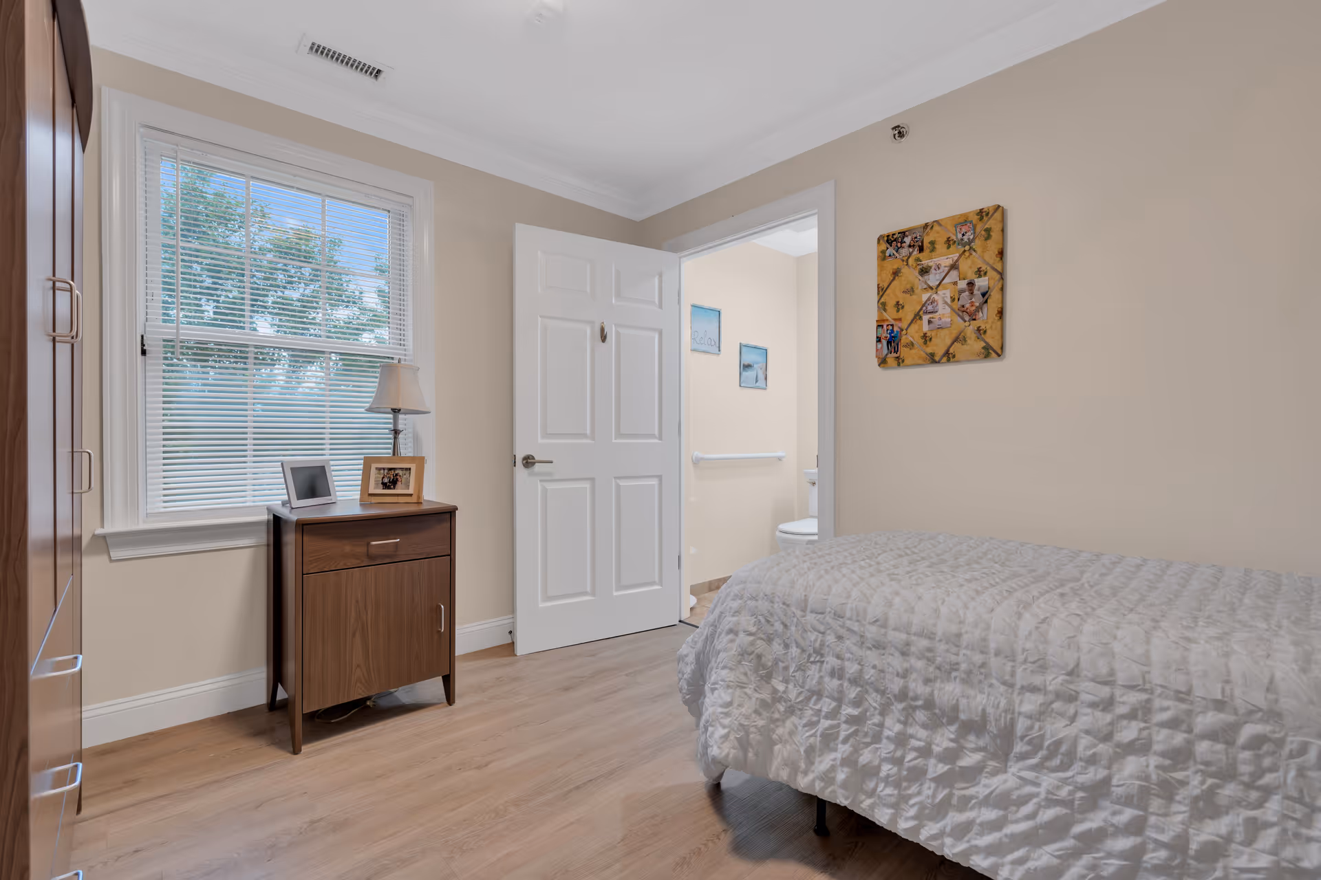 A cozy bedroom with a single bed covered in a white quilted bedspread. There is a wooden nightstand with a lamp and two framed photos next to a window with white blinds. The door to an adjacent bathroom is open, showing a toilet and wall decorations inside. The walls are painted beige and the floor is light wood.