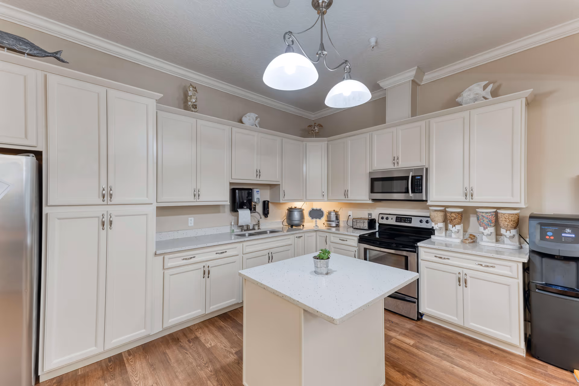 A modern kitchen with white cabinets, stainless steel refrigerator, stove, and microwave. There is a small island in the center with a small potted plant on top. The kitchen has wood flooring and is well-lit with two hanging lights above the island. Various kitchen appliances and cereal dispensers are placed on the countertops.