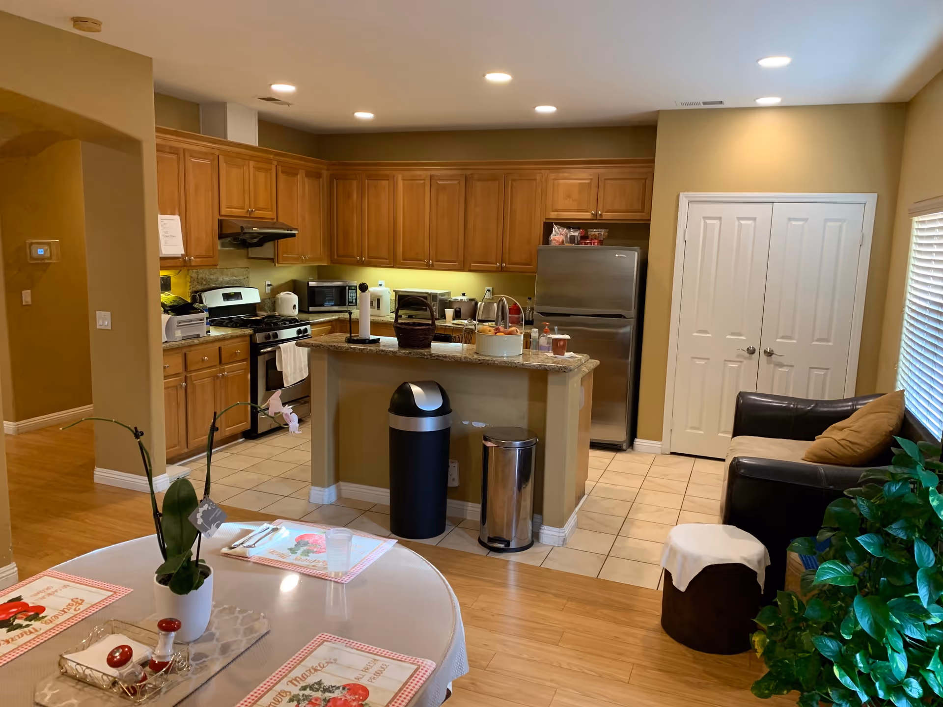 Interior view of a residential kitchen and dining area with wooden cabinets, stainless steel appliances including a refrigerator and stove, a kitchen island with a paper towel holder and basket, a round dining table set with placemats and a potted orchid, a small seating area with a dark leather couch and a small ottoman, and a large green plant near the window with blinds.