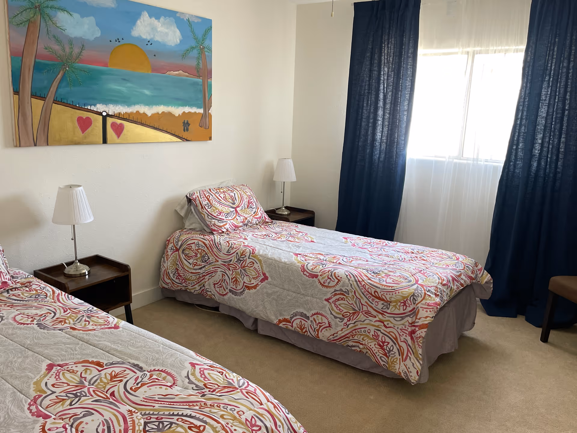 A bedroom with two single beds covered in white bedspreads with pink and orange patterns. Each bed has a matching pillow. There are two small wooden nightstands with white lamps on either side of the beds. A colorful painting of a sunset over the ocean with palm trees and hearts is hung on the wall above one bed. A window with sheer white curtains and dark blue drapes lets in natural light.