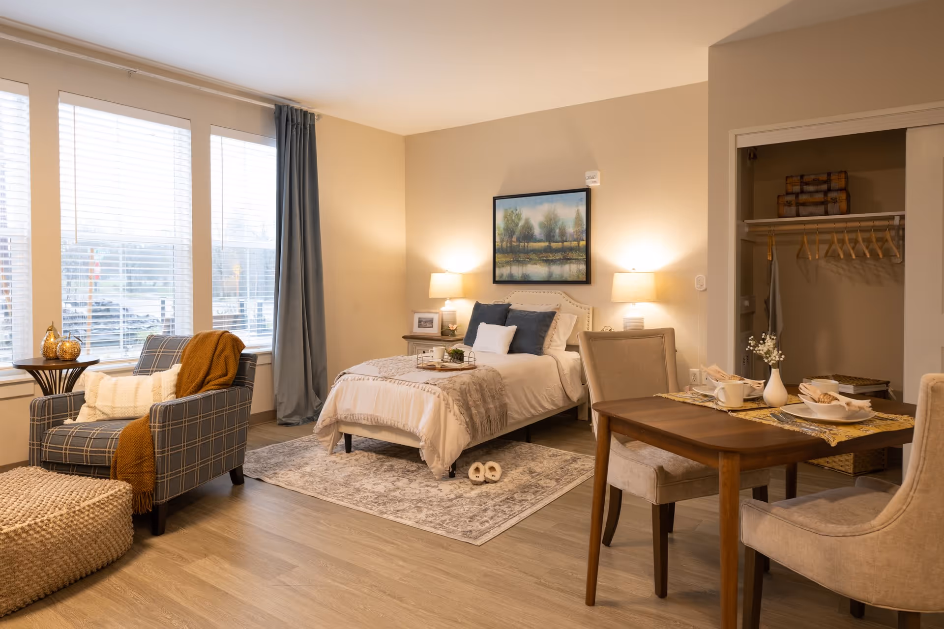 A cozy, well-lit bedroom with a single bed adorned with pillows and a throw blanket. Two bedside tables with lamps flank the bed, and a framed landscape painting hangs above it. To the left, there is a comfortable armchair with a throw blanket and pillows next to a small round table with decorative pumpkins. On the right side, a wooden dining table with two upholstered chairs is set with plates, cups, and a small vase with flowers. An open closet with hangers and storage baskets is visible in the background. Large windows with blinds and curtains allow natural light to fill the room.