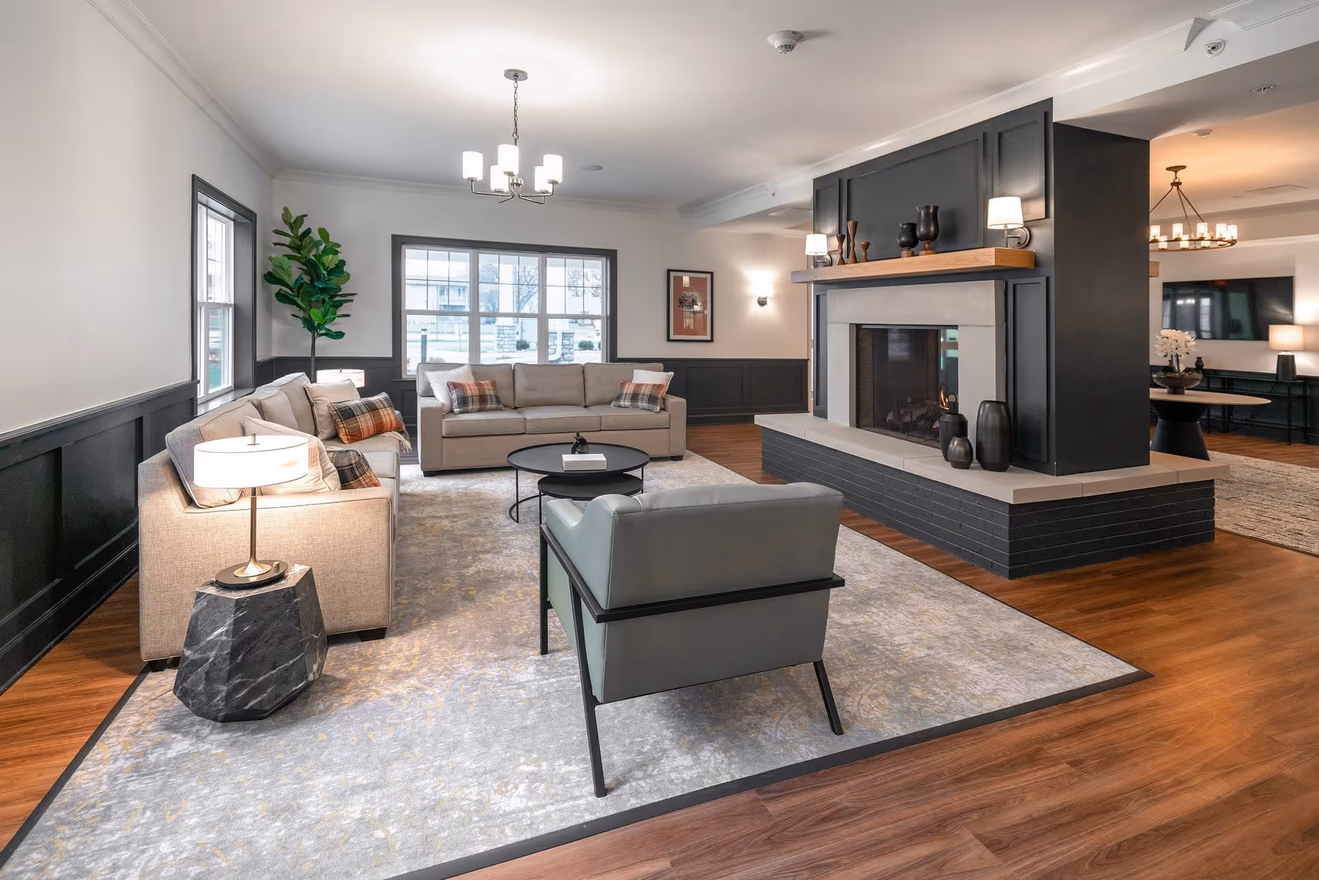 A modern living room with two beige sofas, a gray armchair, a round black coffee table, and a large area rug. The room features a central fireplace with decorative vases on the mantel, wood flooring, and large windows letting in natural light. There are also lamps and wall sconces providing additional lighting.