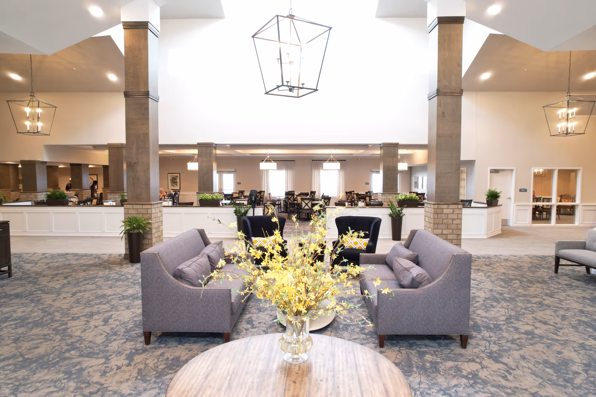 A spacious and well-lit senior living common area featuring two gray sofas facing each other with a round wooden table in the foreground holding a vase of yellow flowers. Behind the sofas are two black armchairs with yellow patterned cushions. The room has high ceilings with modern hanging light fixtures and several columns. In the background, there are multiple dining tables and chairs arranged neatly, with large windows allowing natural light to fill the space.