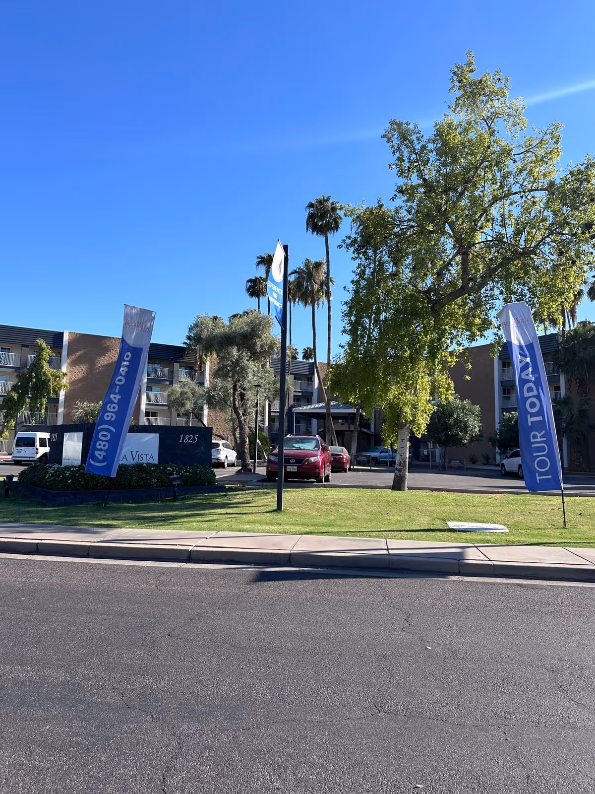 Front exterior of Bella Vista Senior Living with flags, a lawn, and parked cars under a clear blue sky.
