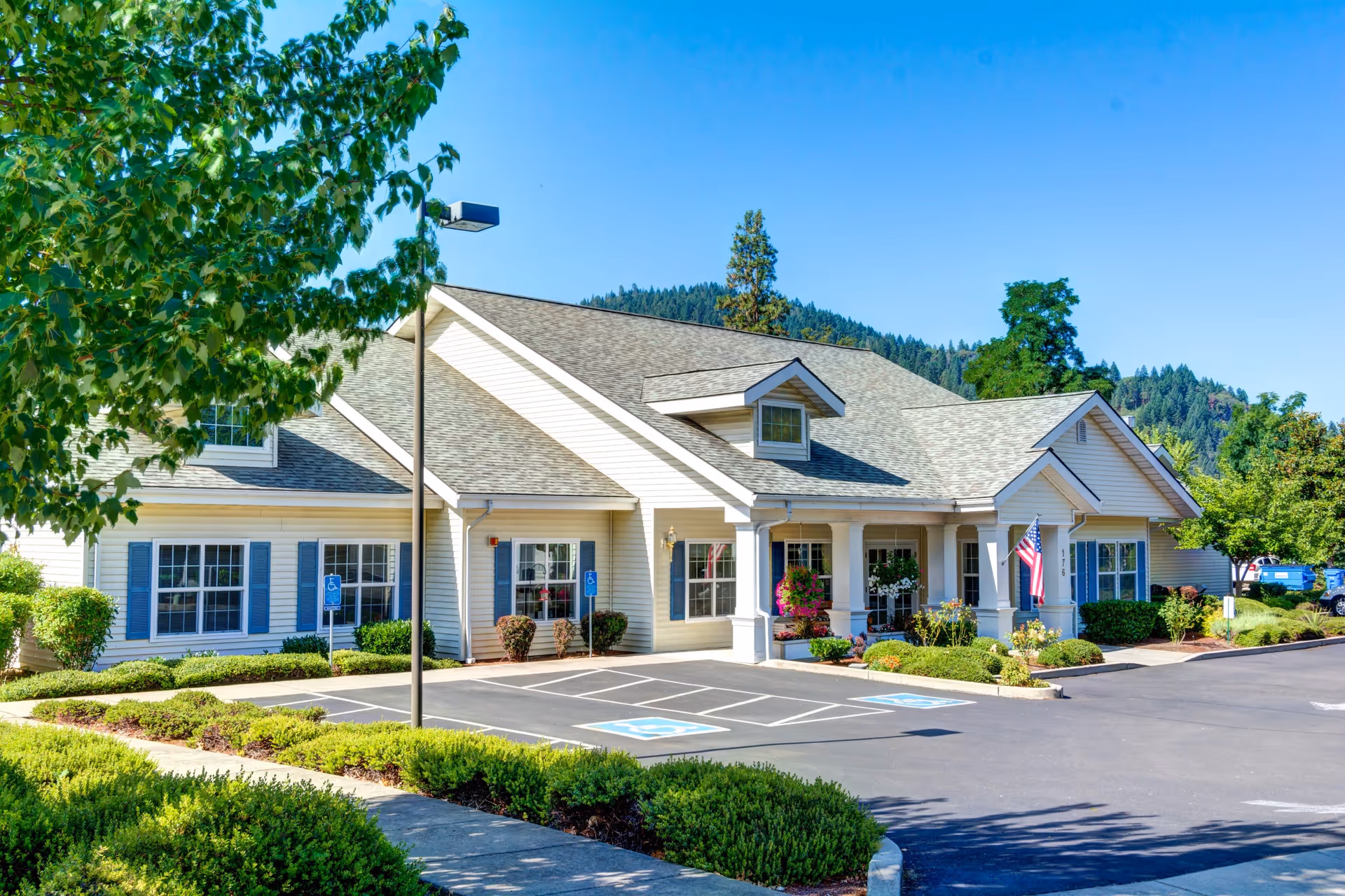 Exterior view of Morrow Heights Assisted Living facility showing a single-story building with a gray roof, white siding, blue window shutters, and a covered entrance with columns. The parking lot in front has designated handicapped parking spaces, and there are well-maintained bushes and trees around the building under a clear blue sky.