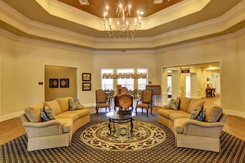 Well-lit communal sitting area with sofas and armchairs arranged around a central table and chandelier, with a dining area visible in the background.