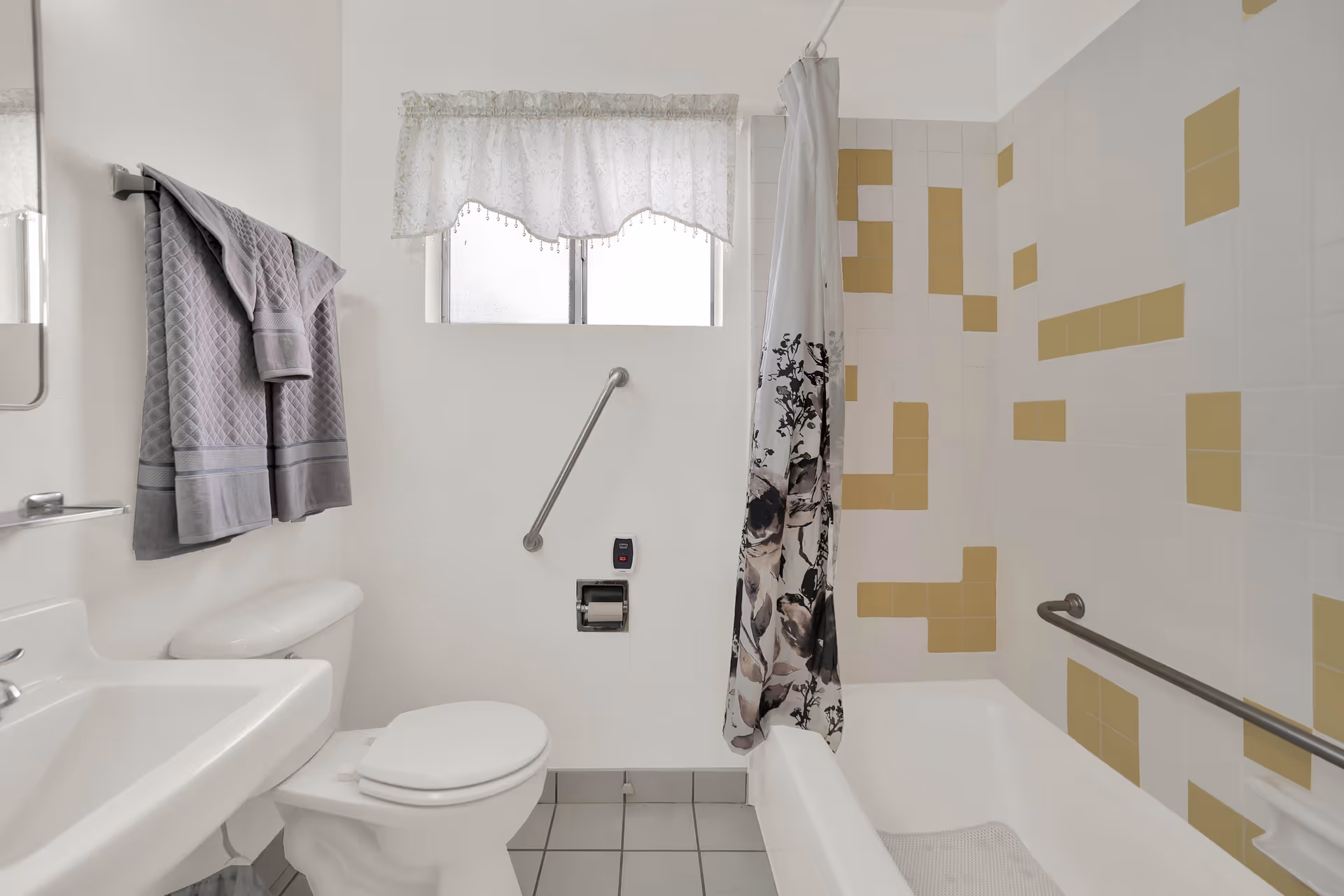 Bright accessible bathroom with a toilet and sink, bathtub/shower with a floral curtain, grab bars, and patterned wall tiles.