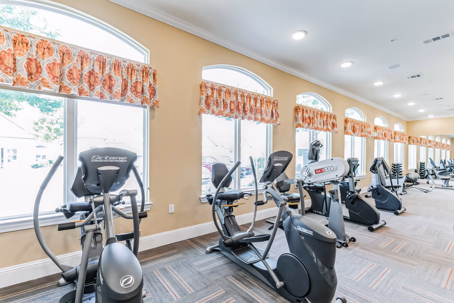 A bright fitness room with multiple exercise machines including stationary bikes and elliptical trainers lined up along a wall with large windows covered by patterned valances. The room has beige walls, recessed lighting, and a carpeted floor.